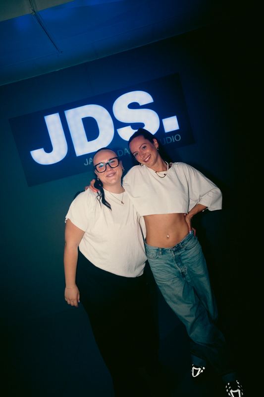 Zwei Trainerinnen lachen Arm in Arm unter dem JDS-Leuchtschild